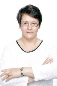 Portrait of Elna Esterhuizen, wearing glasses and a white top, with arms crossed and a neutral expression.