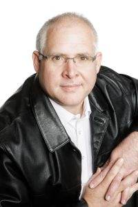 Man with glasses, wearing a black leather jacket and white shirt, looking into the camera. Daniel Esterhuizen.