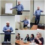 A man in a blue shirt gives a presentation to a group of attentive women in a meeting room with a projector and flipchart.