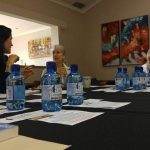 Three women are seated at a table with water bottles, documents, and mugs, engaged in conversation in a room with colorful artwork.