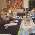 Three women sitting at a table, writing on papers, with colorful artwork and an orchid centerpiece in a cozy room.