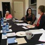 Four women sit around a conference table with documents, notebooks, and bottled water, engaged in discussion.