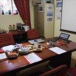A conference table setup with snacks, papers, and a laptop; coffee station and window with red curtains in the background.