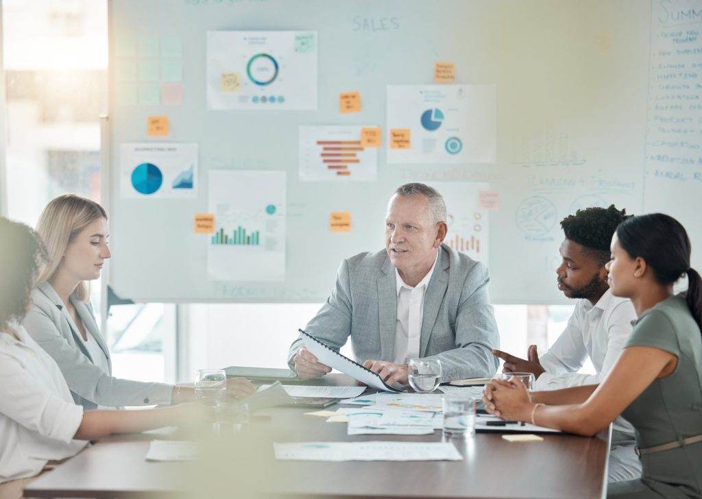 A group discussion during an executive coaching session with charts and data on a whiteboard in the background.
