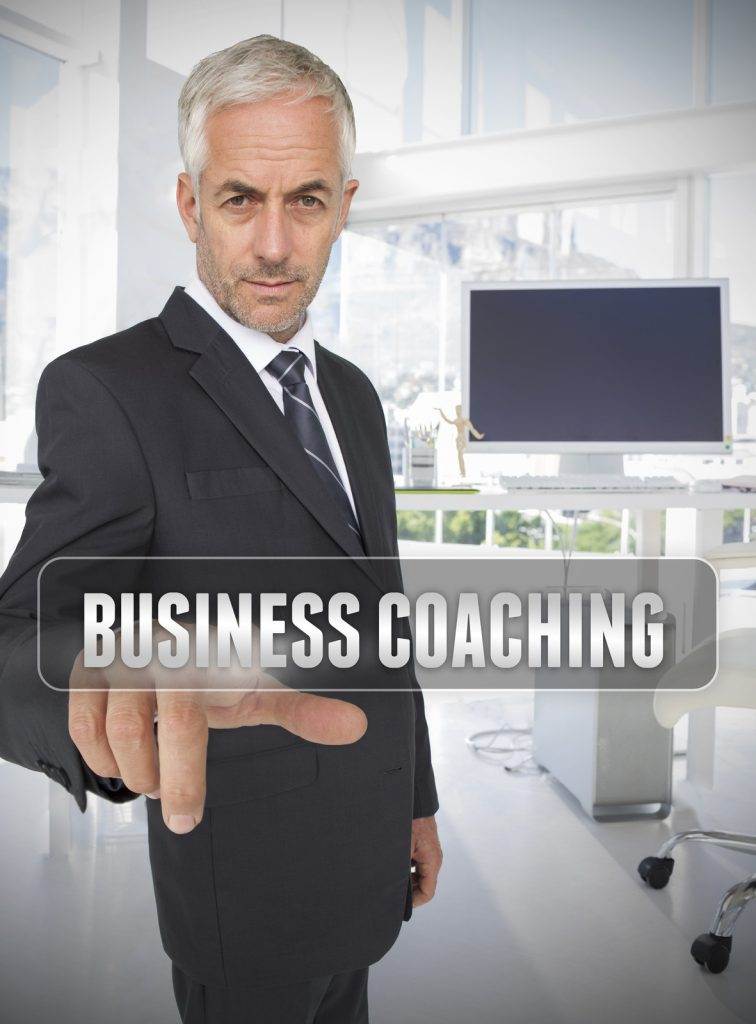 A man in a suit pointing at a translucent "Business Coaching" text overlay in a modern office setting.
