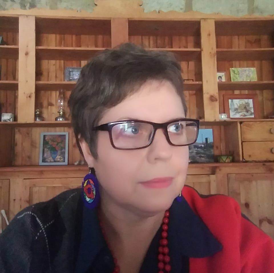 Marna van den Berg wearing red and blue earrings and glasses, in front of a wooden bookshelf with various decorations.