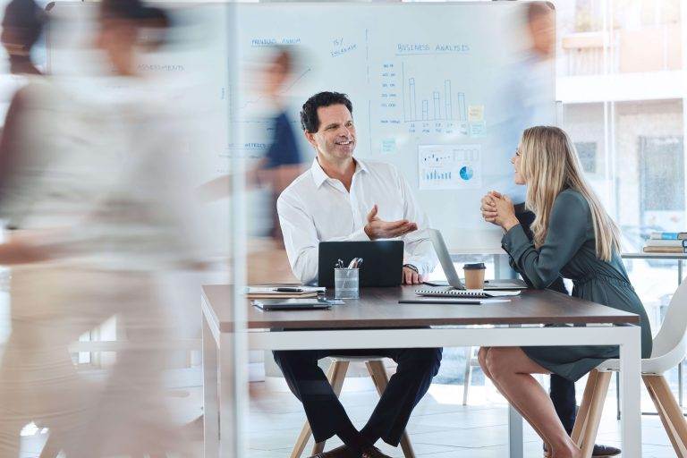 An executive coaching session with a man and woman seated at a table, discussing business charts on a whiteboard.