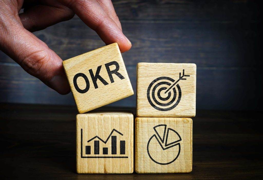 A hand stacking wooden blocks labeled OKRs, symbolizing objectives, key results, and delivering strategic goals.