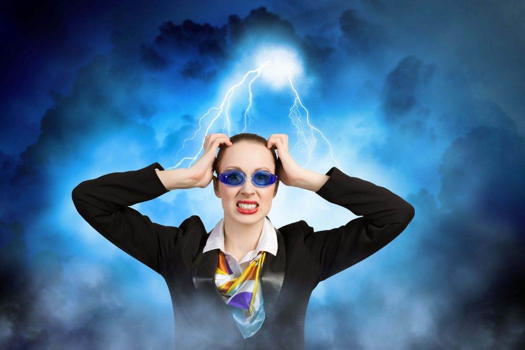 Person in a suit and colorful scarf, wearing blue goggles, holds head with a grimace, background of storm clouds and lightning.
