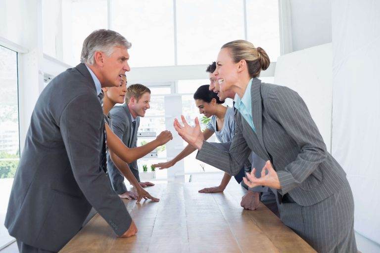 Two groups of businesspeople in a heated discussion, facing each other across a conference table in a bright office.
