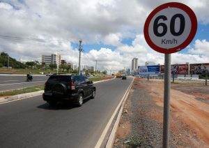 Urban road with a 60 km/h speed limit sign, vehicles, billboard advertisements, buildings, and a partly cloudy sky.