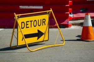 Yellow "Detour" sign with a black arrow pointing right, placed on a road near a traffic cone and red barriers.