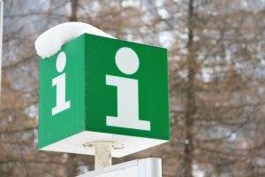 Green information sign with white 'i' symbols, covered with snow, in front of a snowy blurred forest background.