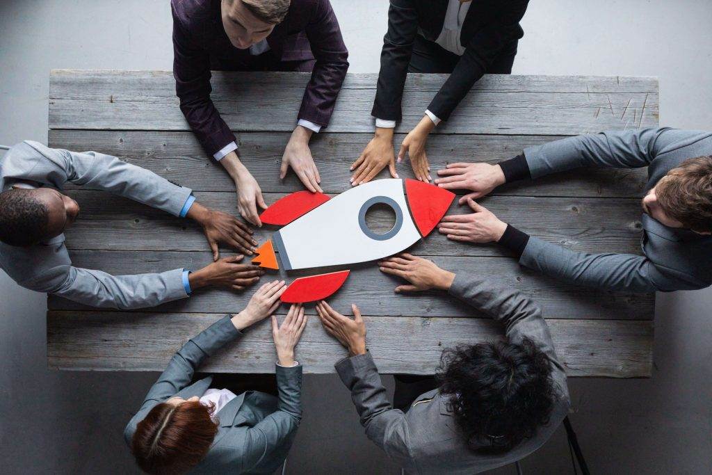 Diverse team collaborates around a table, placing hands on a rocket model, illustrating team development interventions.