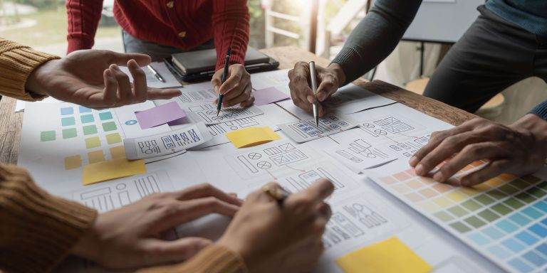 Hands of diverse individuals collaborating on design sketches and color charts on a wooden table, engaged in creative planning.