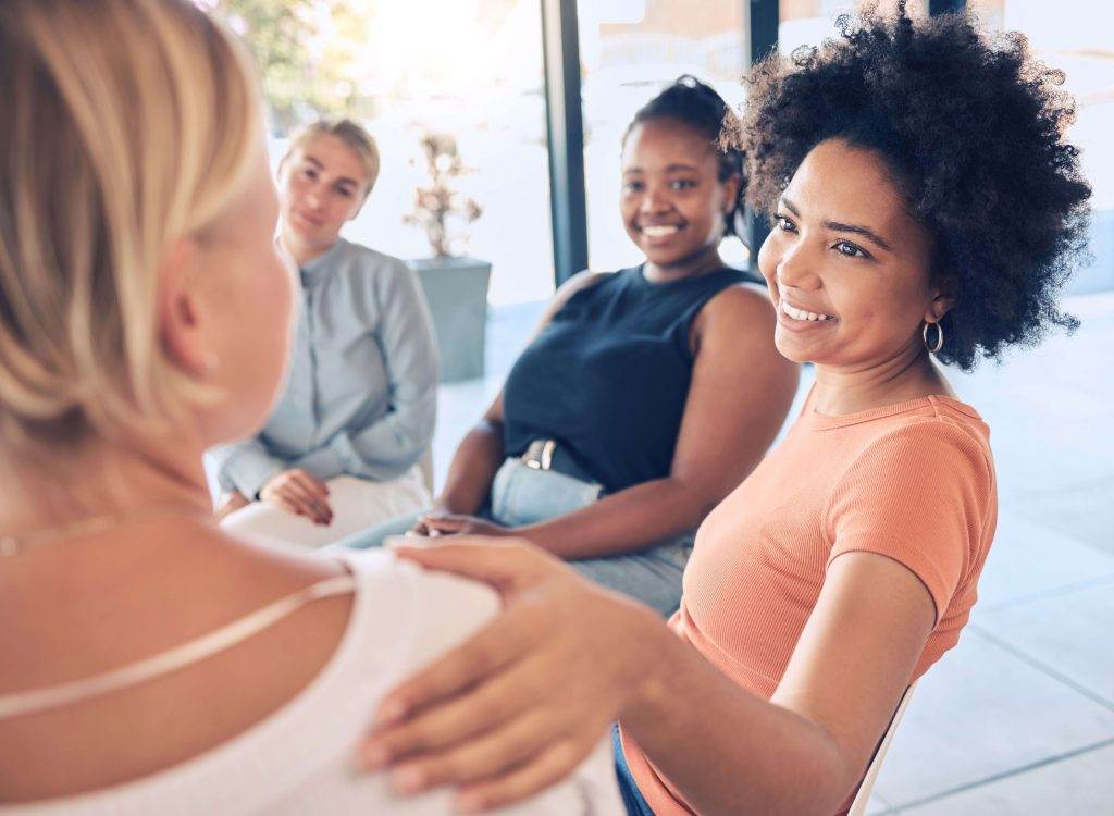 Four women sitting in a circle during a support group session, one woman comforting another by placing a hand on her shoulder.