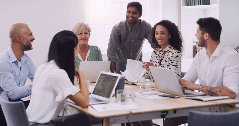 A diverse group of six colleagues in a meeting room smiling and discussing work, with laptops and documents on the table.
