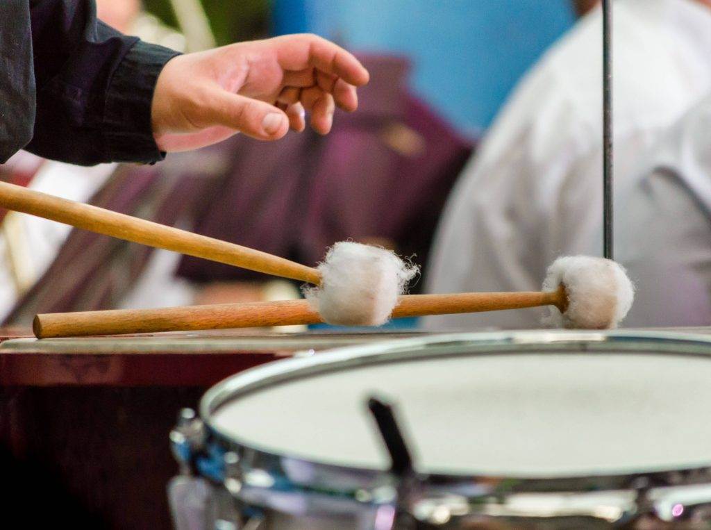 A hand holding drumsticks with mallets, playing a snare drum, with musicians blurred in the background.