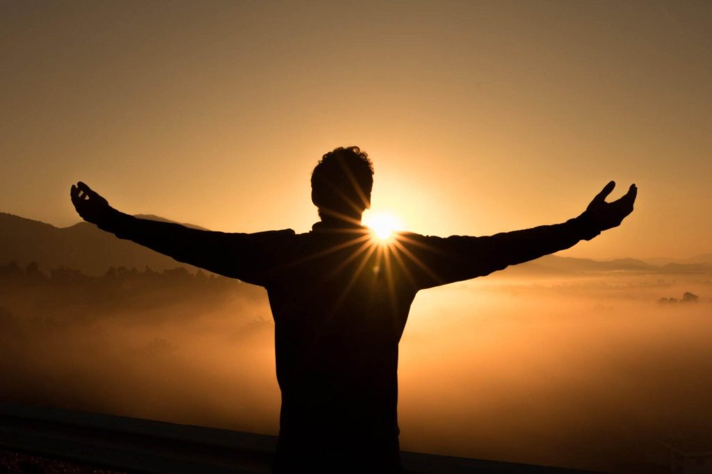 Silhouette of a person with arms outstretched, standing before a sunrise over a misty landscape.