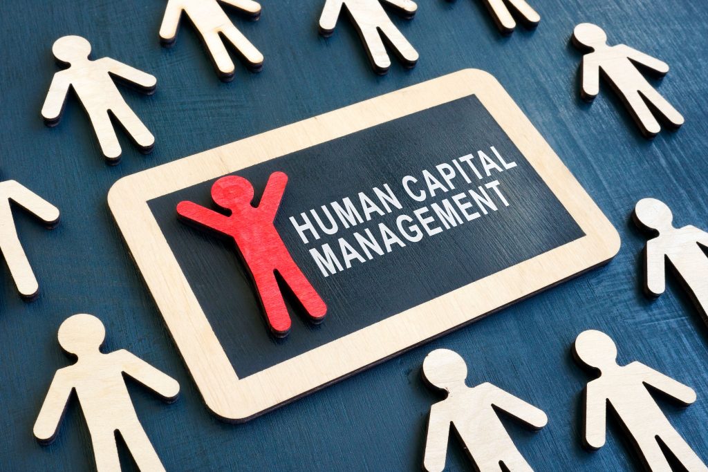 Wooden figures surround a plaque reading "Human Capital Management," representing HR consulting.