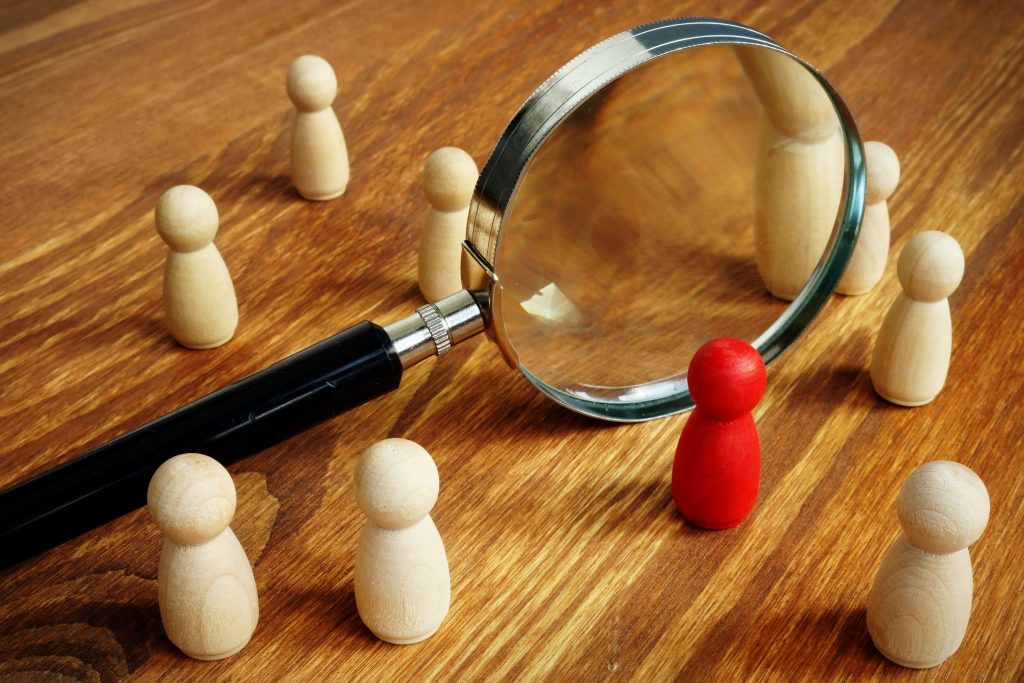 Magnifying glass focusing on a red wooden figure among beige ones, symbolizing job profiling and candidate selection.