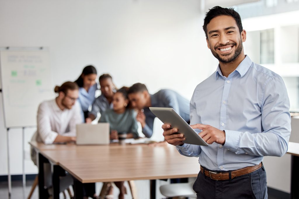 Smiling man with a tablet in front of a team discussing project management in a meeting room.