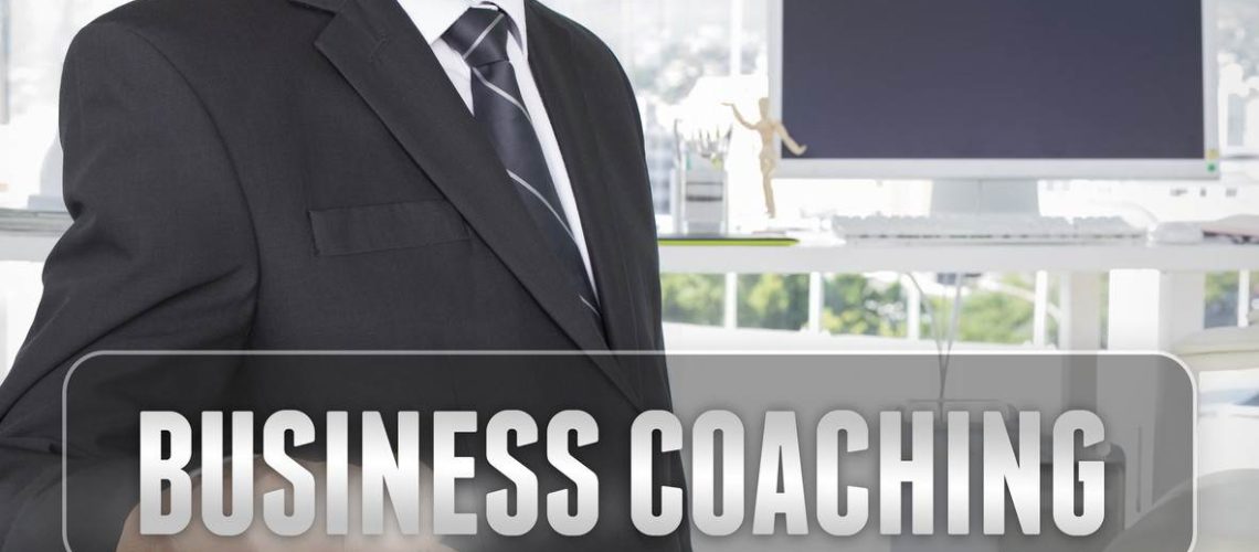 A man in a suit pointing at a translucent "Business Coaching" text overlay in a modern office setting.