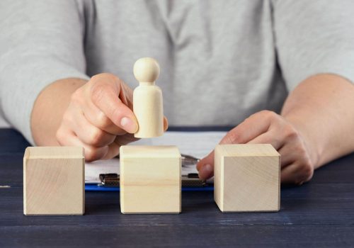 A person arranges a wooden figure between three wooden blocks on a table, with a clipboard in the background.