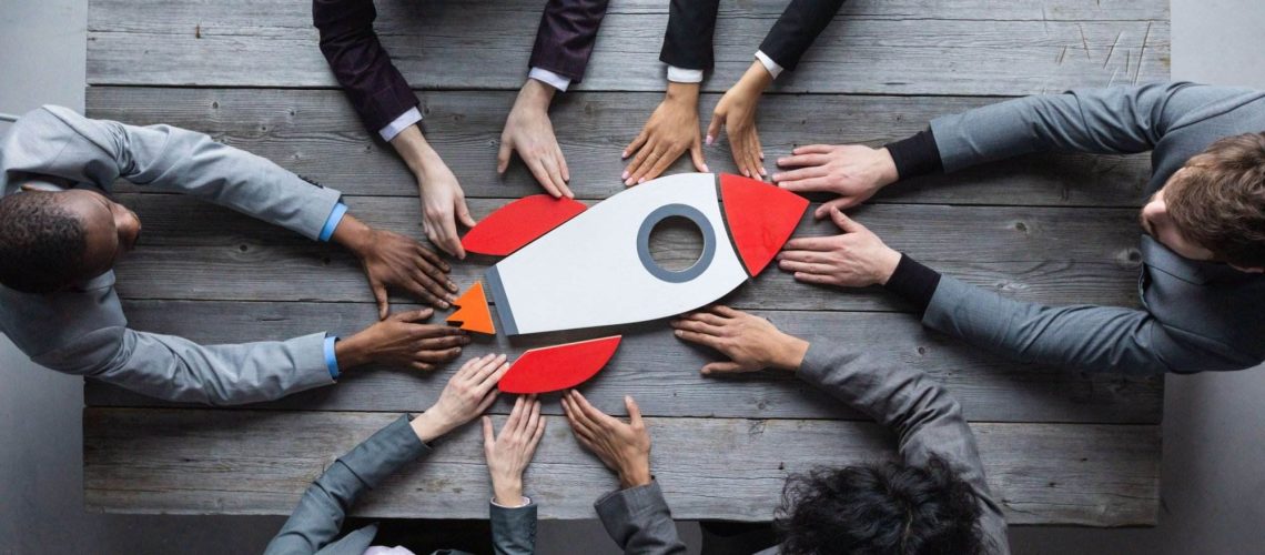Diverse team collaborates around a table, placing hands on a rocket model, illustrating team development interventions.
