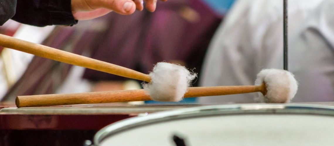 A hand holding drumsticks with mallets, playing a snare drum, with musicians blurred in the background.
