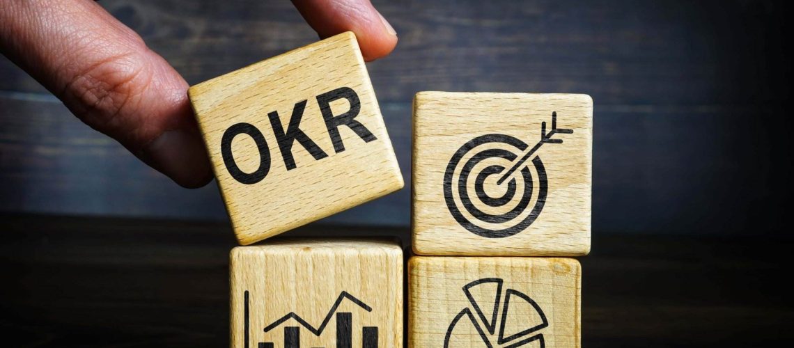 A hand stacking wooden blocks labeled OKRs, symbolizing objectives, key results, and delivering strategic goals.